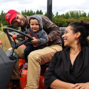 Man On A Tractor With A Young Child On His Lap, A Woman Looks At Them Smiling In The Forground